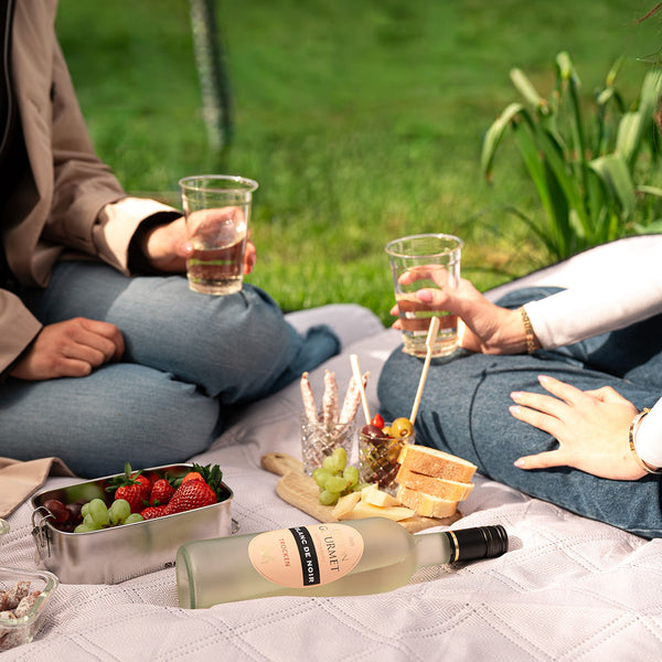 Zwei Personen genießen ein Picknick im Gras mit Edition Gourmet Blanc de Noir, Obst und Brot