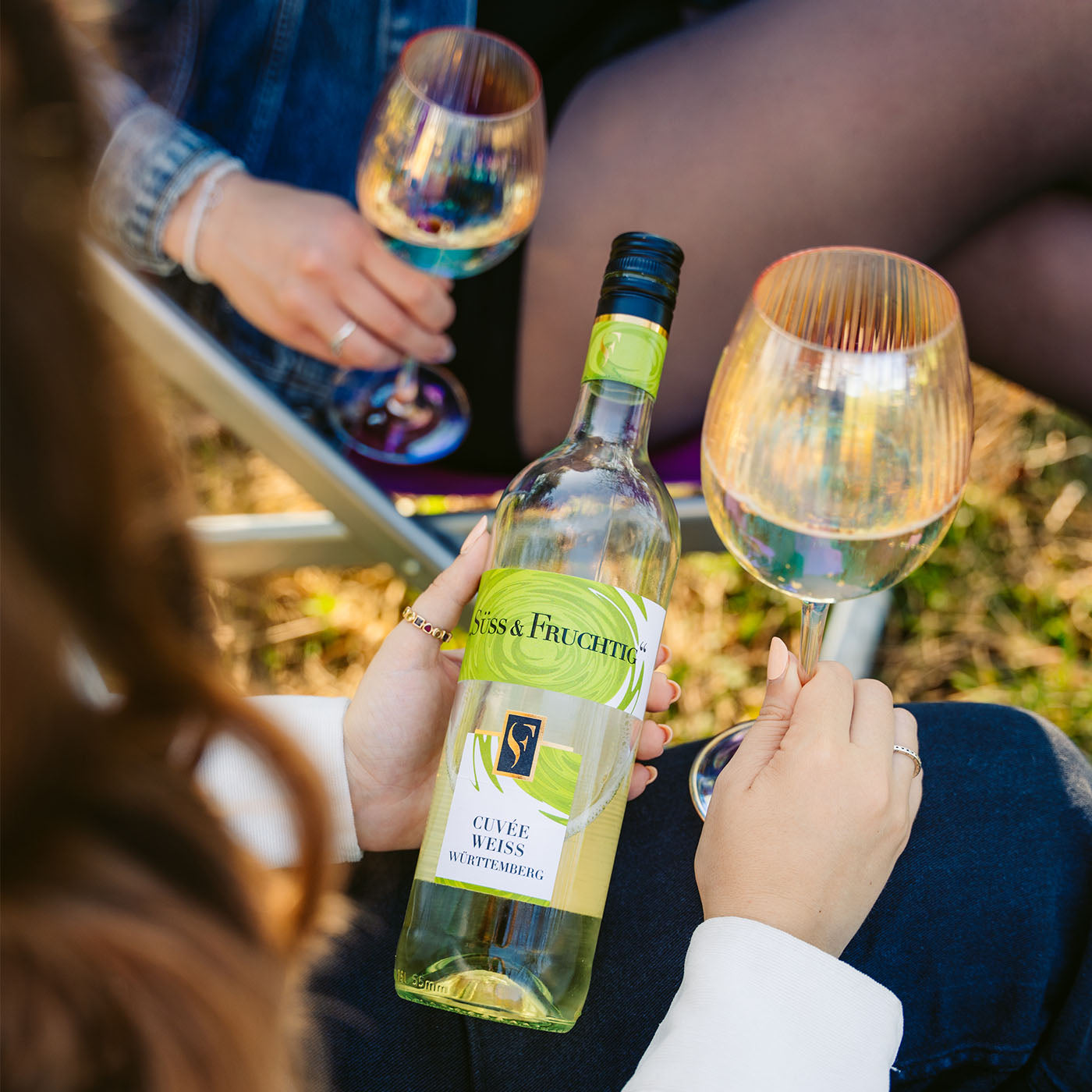 Flasche Süss & Fruchtig Cuvée Weiss wird zwischen gefüllten Weingläsern im Freien gehalten